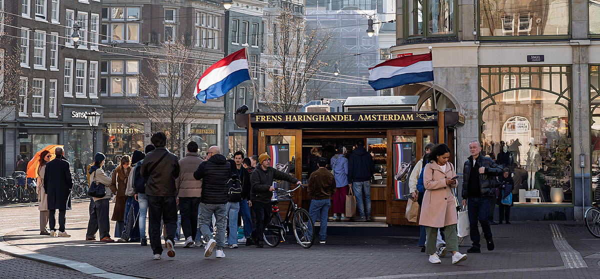 Gebiedsopgaven Amsterdam, Area Challenges, Amsterdam street with people near a traditional Dutch herring stall, flags waving
